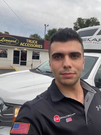 Homme portant un polo noir avec logo Spacekap debout devant un camion blanc stationné près d'un magasin Leonard Truck Accessories