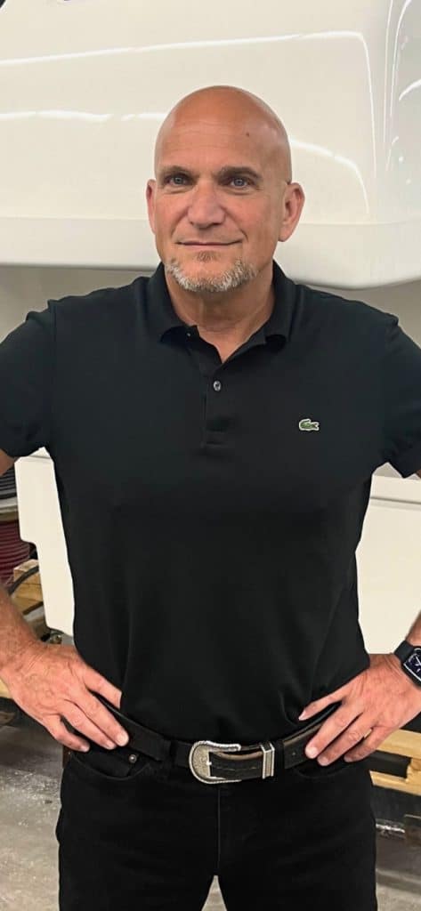 Homme chauve portant un polo noir avec logo crocodile et une ceinture noire, posant avec les mains sur les hanches
