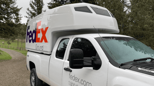 Camionnette blanche avec logo FedEx Ground stationnée sur un chemin en gravier avec des arbres en arrière-plan